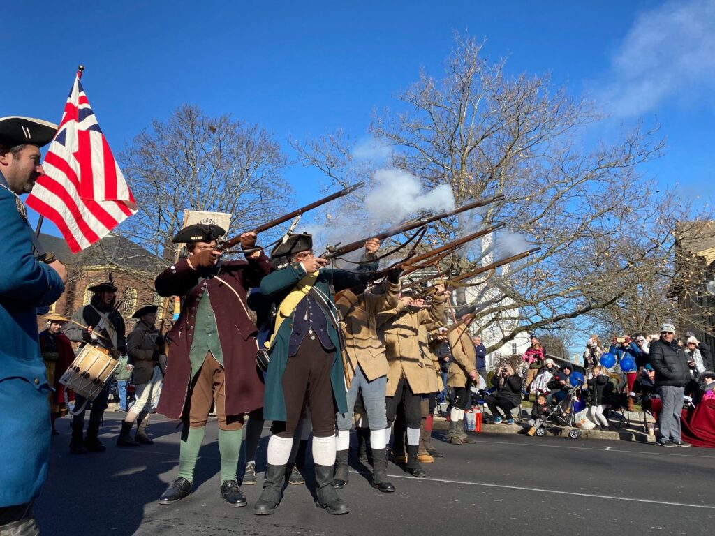 Stow Minutemen firing a rifle volley. The flag, with a British Union Flag in the upper left corner, combined with 13 red and white stripes to represent the colonies, was called the Grand Union Flag, used by Washington's Army in 1776.